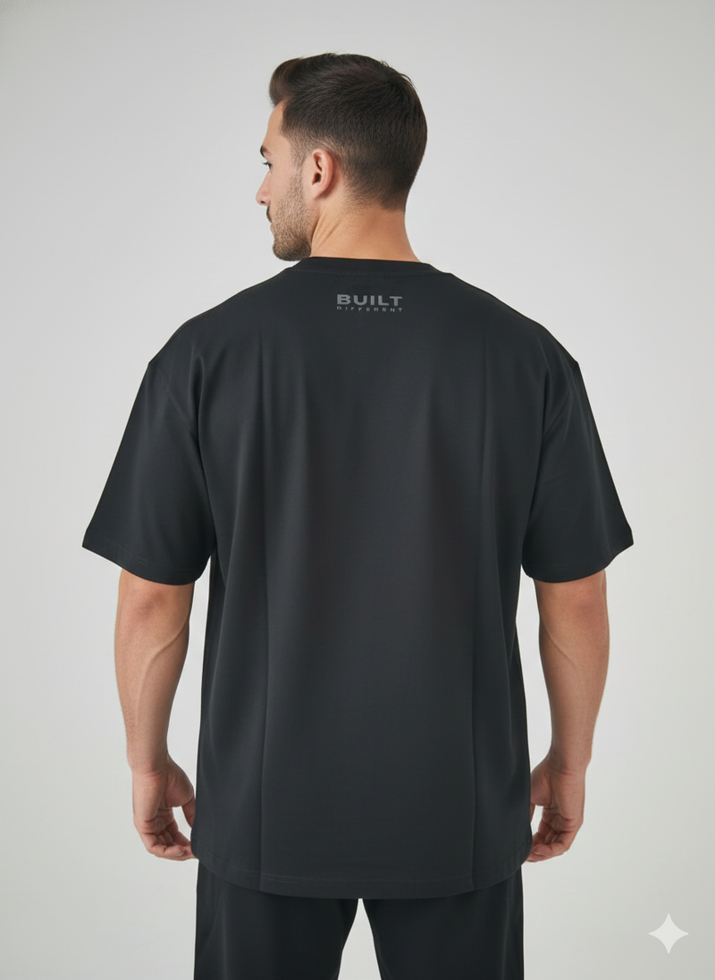 A man faces away from the camera, wearing a black BDHQ: Oversized T-Shirt (180GSM) – Lightweight Drop-Shoulder Cut by Men's Gym Clothes AU, with "BUILT" in gray lettering on the back near the neckline. He also wears black pants against a plain light background.