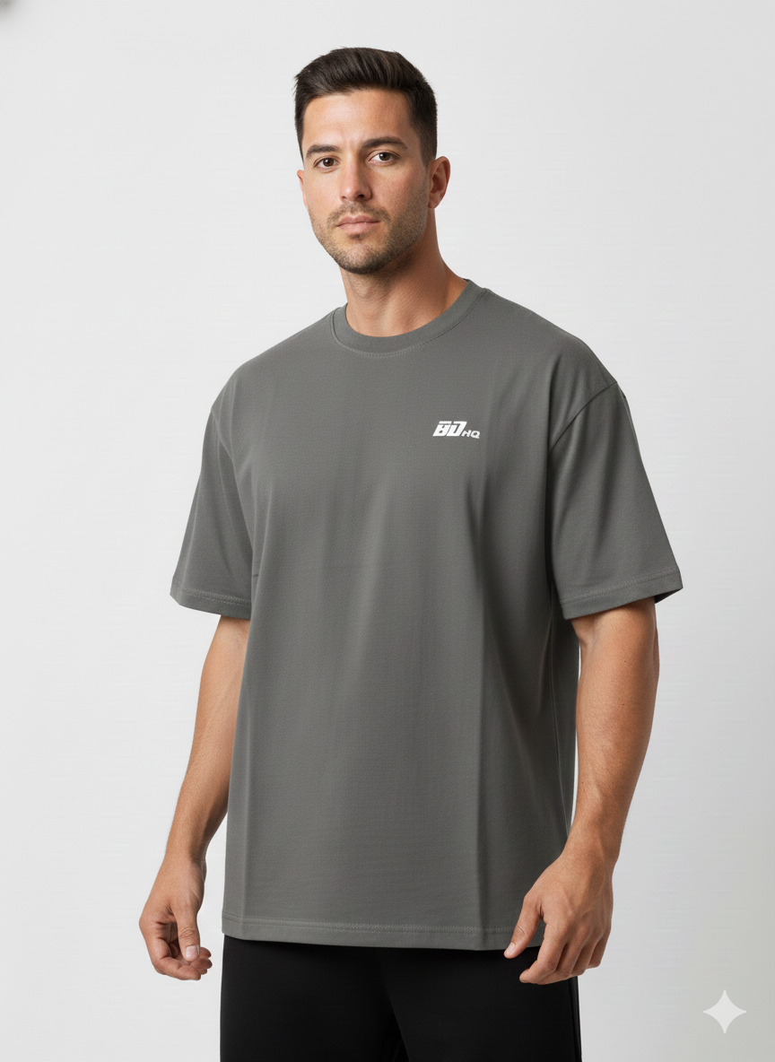 A man wears the BDHQ: Oversized T-Shirt (180GSM) from Men's Gym Clothes AU—a loose-fitting, dark gray, drop-shoulder cotton tee with a small white chest logo—standing against a white background and looking at the camera neutrally.