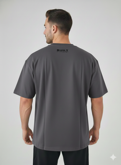 A man faces away from the camera wearing the Men's Gym Clothes AU BDHQ Oversized T-Shirt (180GSM), lightweight with a drop-shoulder cut, in dark gray. "BUILT" and small text are printed on the upper back against a light background.